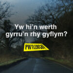 A driver's view of the road with the text "Yw hi'n werth gyrru'n rhy gyflym?"